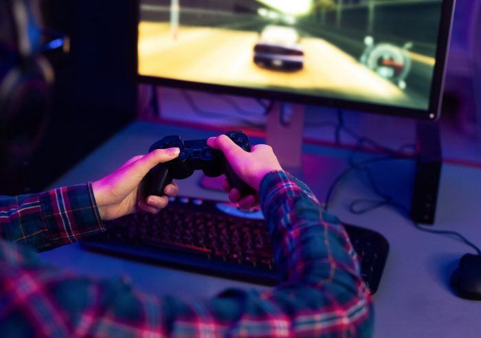 This image shows a gamer playing a racing video game at their computer desk. The gamer is wearing a plaid shirt and using a black video game controller. The computer monitor is displaying racing game